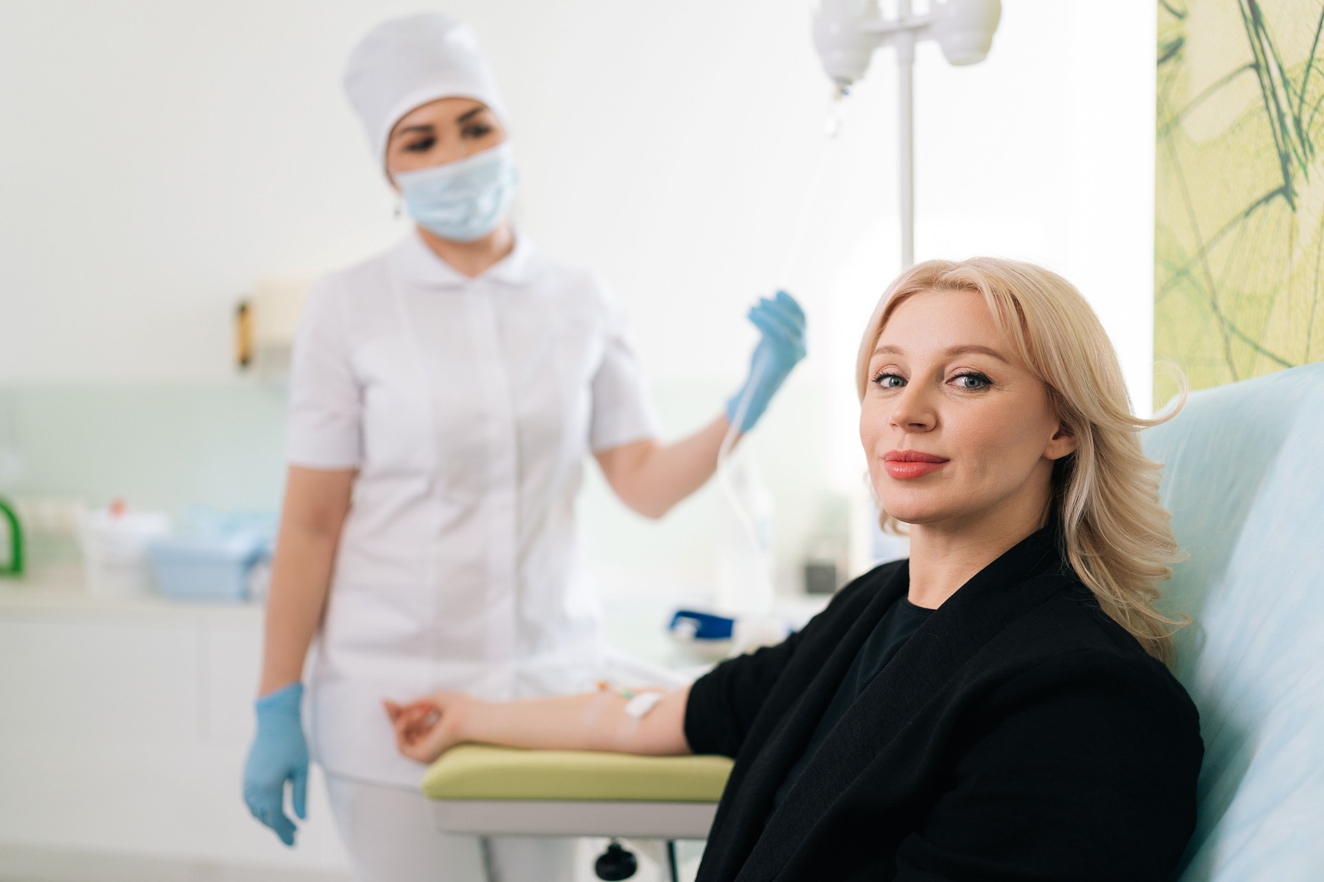 Portrait of blonde woman sitting on medical chair and getting intravenous vitamin therapy looking at camera. Clinic nurse in uniform standing on blurred background.